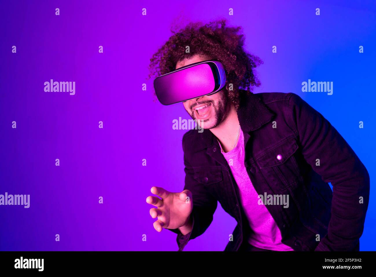 CONCETTO DI METAVERSE. Sorprende il giovane uomo con capelli afroche toccano l'aria e sorridono durante l'esperienza VR. Scatto orizzontale in studio. Viola blu Foto Stock