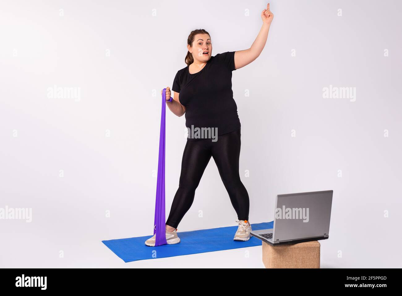 Attività domestica e formazione, corso di fitness online. Donna bionda sovrappeso che fa esercizi di stretching, banda di resistenza violetta che guarda video guida Foto Stock
