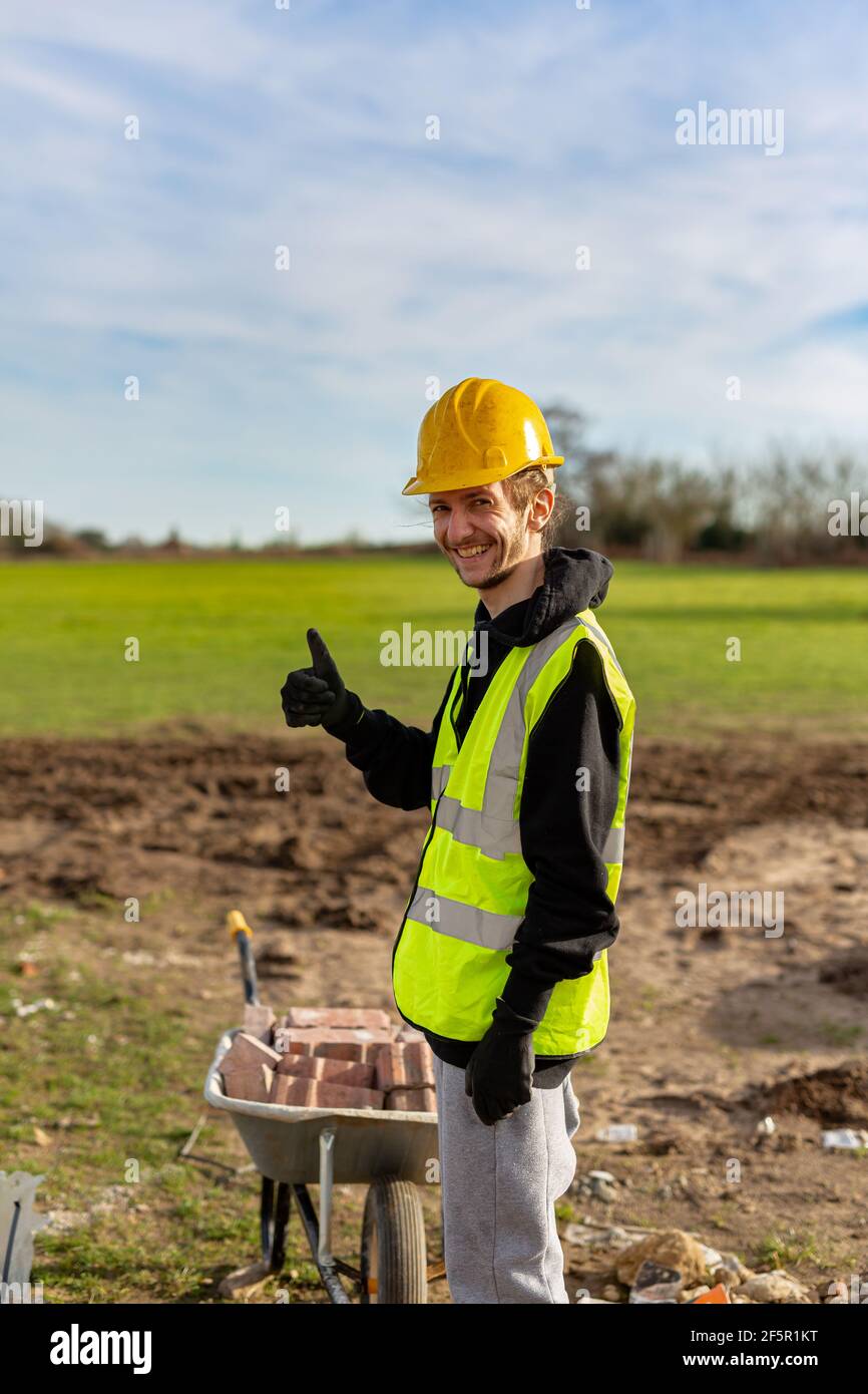 Un giovane adulto maschio costruttore che indossa un gilet ad alta visibilità e elmetto mentre dà un pollice in su Foto Stock