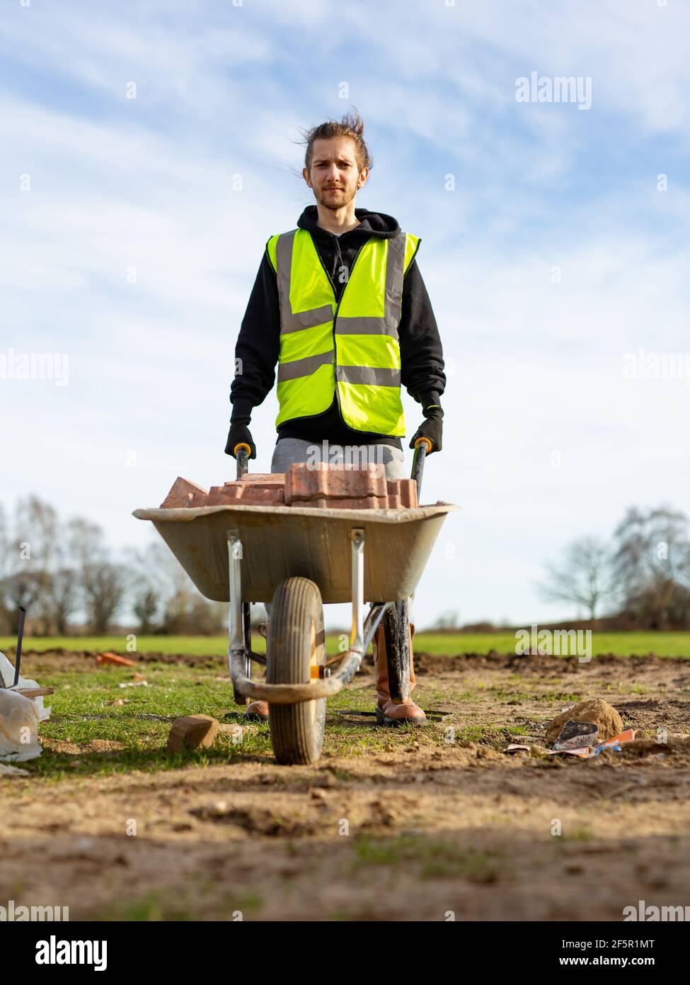 Un giovane adulto maschio costruttore che indossa un gilet ad alta visibilità e elmetto che spinge una carriola piena di mattoni mentre su un cantiere Foto Stock