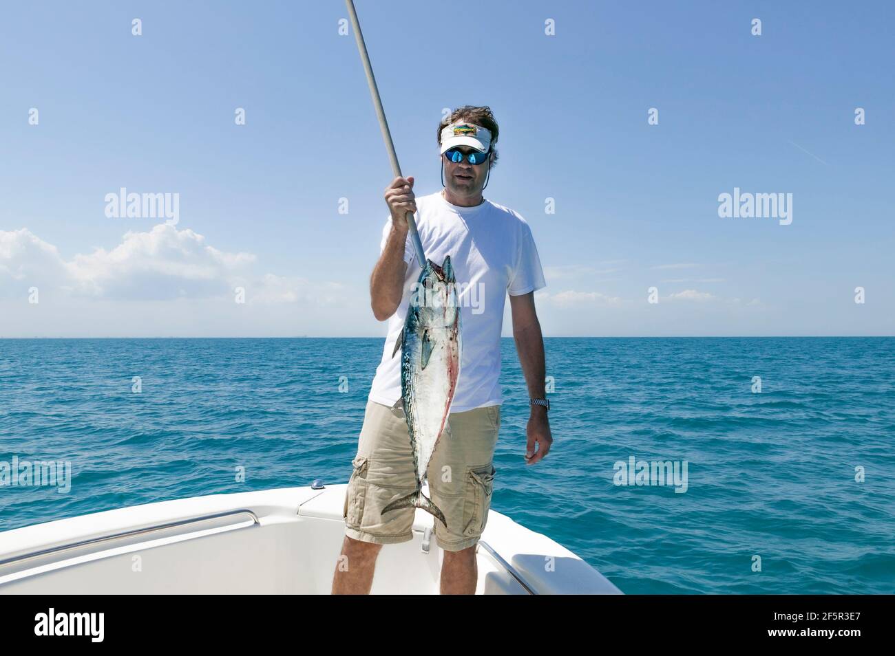 Pescatore della Florida che tiene un piccolo Tunny (Euthynnus alletteratus) sospeso da un gancio di pula mentre è in piedi nella prua Di una barca con l'Atlantico Oce Foto Stock