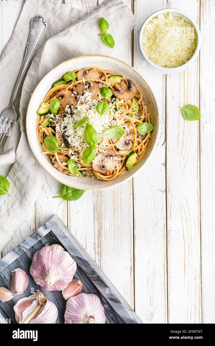 Sani spaghetti di grano integrale con aglio e zucchine di funghi saltati, conditi con formaggio grattugiato e foglie di basilico su rustico fondo di legno Foto Stock