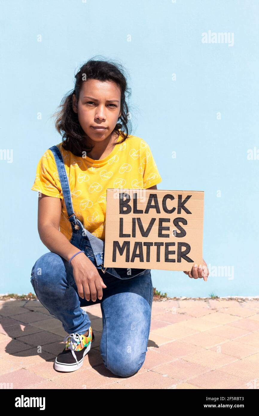 Ritratto di donna latino-americana con atteggiamento serio che tiene un banner con slogan Black Lives materia. Si sta inginocchiando accanto a una parete blu chiaro. Centro benessere Foto Stock