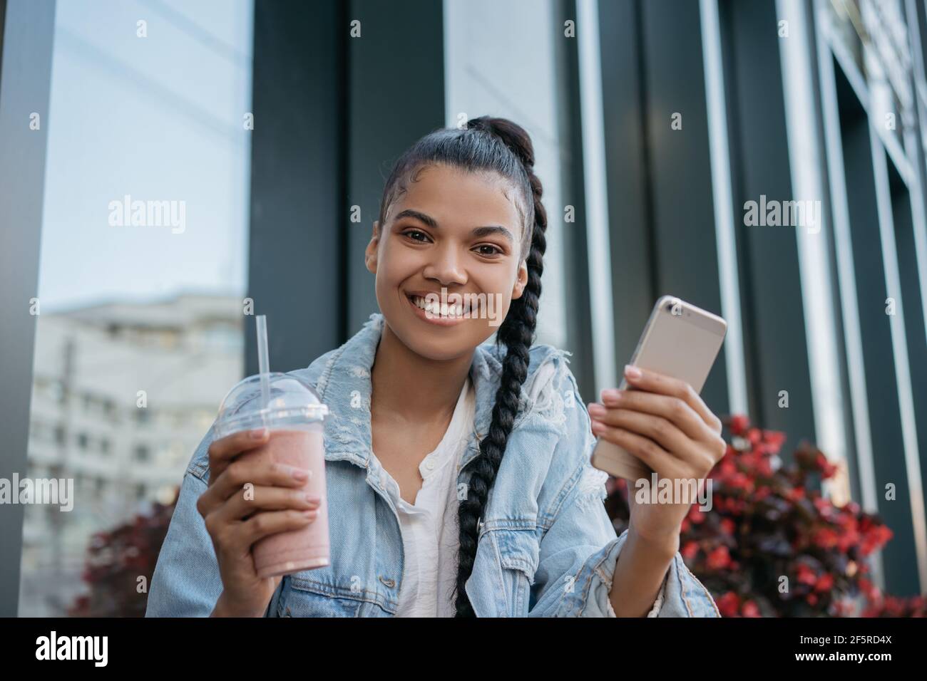 Bella donna afroamericana che tiene il vetro con frullato e smartphone, guardando la fotocamera, sorridendo Foto Stock