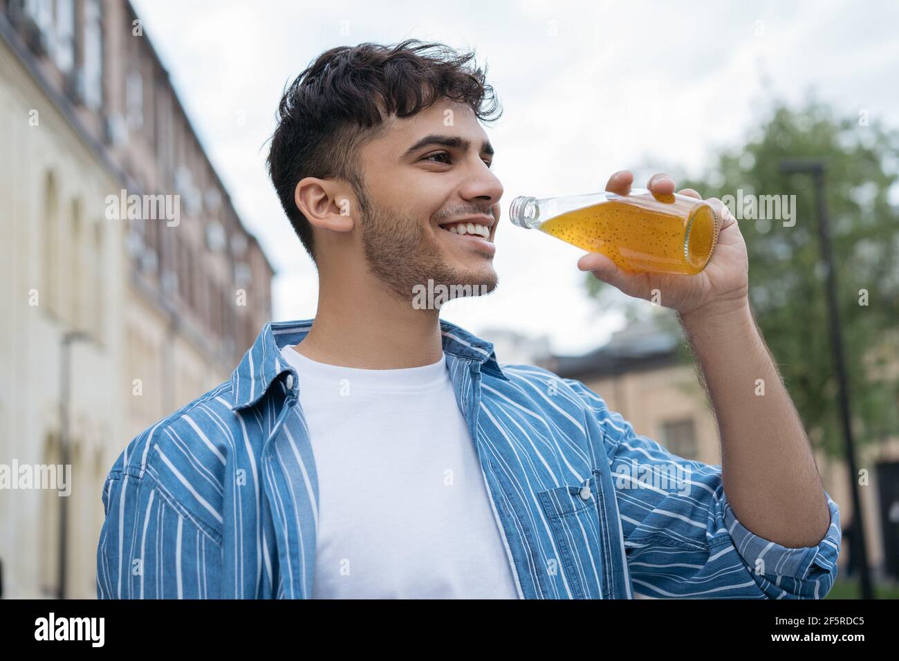 Giovane uomo indiano di stile che tiene il bicchiere con bevande, a piedi per strada, Ritratto di ragazzo asiatico felice indossando abiti casual, bere succo all'aperto Foto Stock