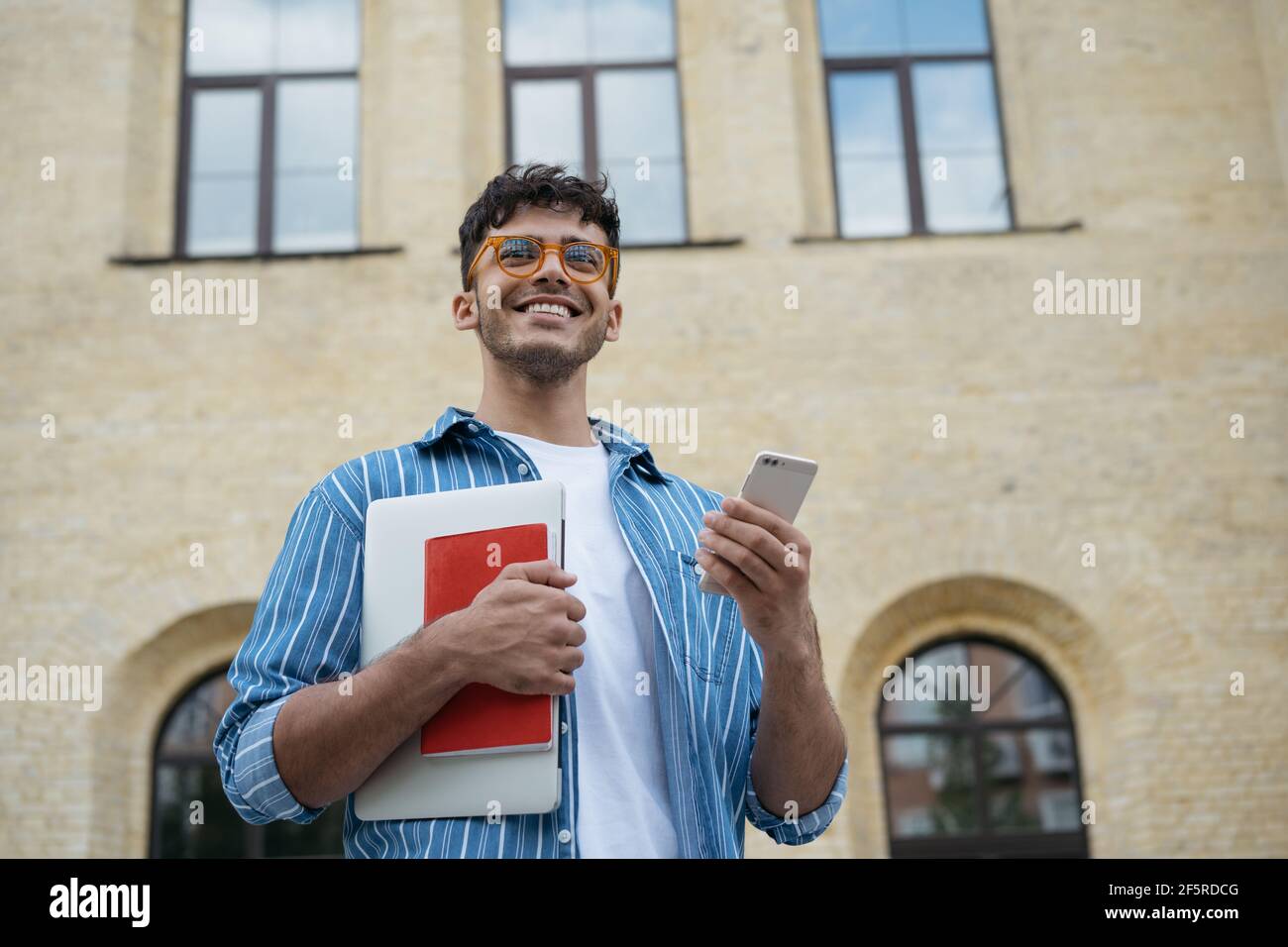 Ritratto di giovane bello studente indiano che tiene portatile, libro, telefono cellulare, a piedi per l'università. Sorridente uomo d'affari asiatico utilizzando lo smartphone Foto Stock