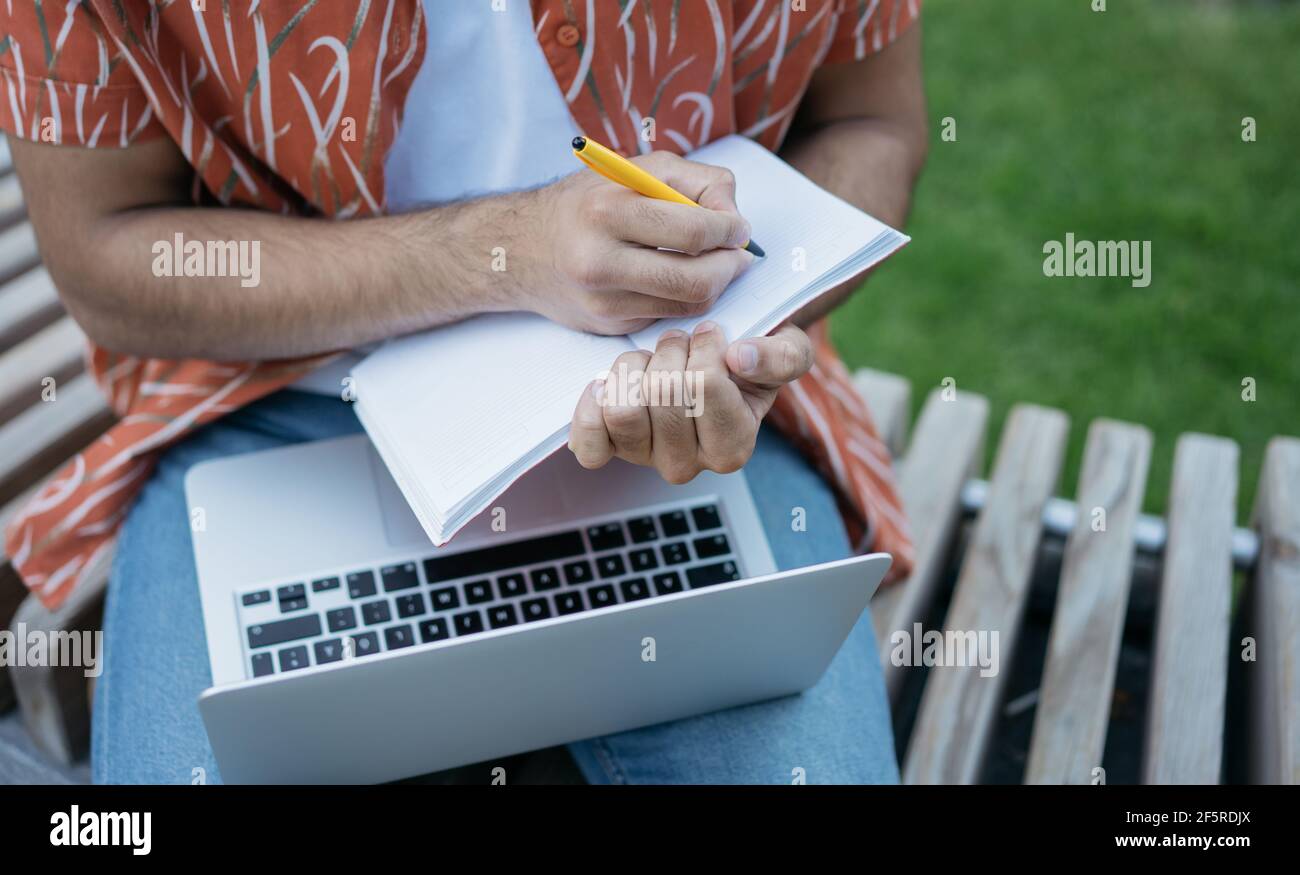 Primo piano di mani maschili prendendo appunti sul notebook, utilizzando il computer portatile, progetto di pianificazione. Uomo d'affari che lavora in linea all'aperto, fuoco sulla mano Foto Stock