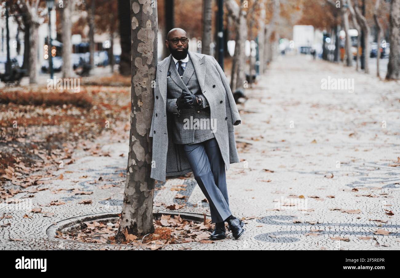 Un cappero nero imprenditore senior in occhiali e un elegante vestito con un costume su misura e un cappotto, si appoggia contro un albero su un autunno b Foto Stock