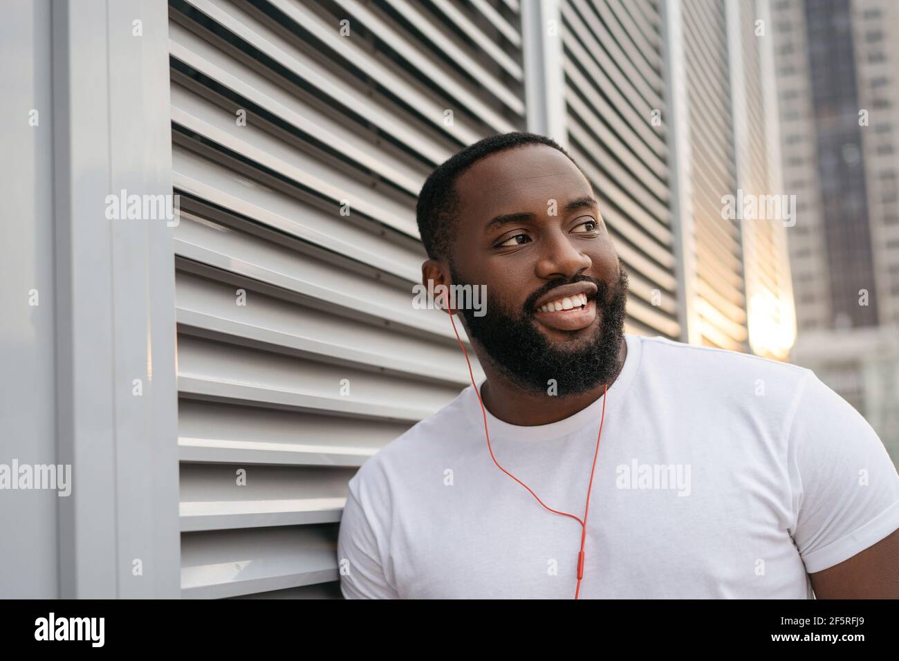 Autentico ritratto di bell'afroamericano che posa per immagini, sorridente, in piedi per strada. Ragazzo con un buon porticato che ascolta musica all'aperto Foto Stock