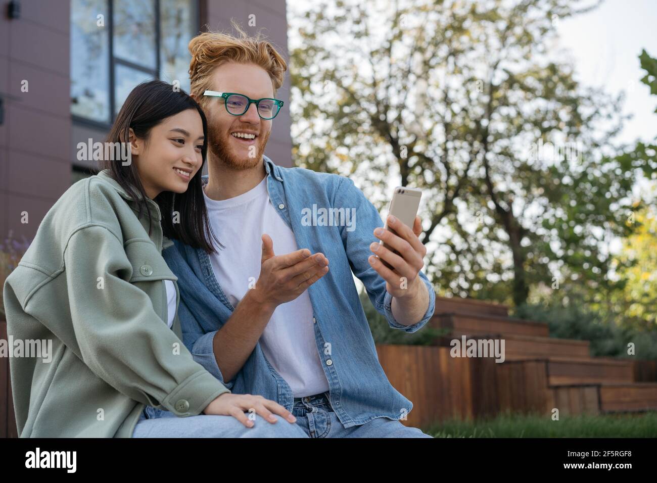 Due amici sorridenti che usano il telefono cellulare per fare acquisti online. Bella donna asiatica e bell'uomo caucasico comunicazione insieme, guardando video Foto Stock