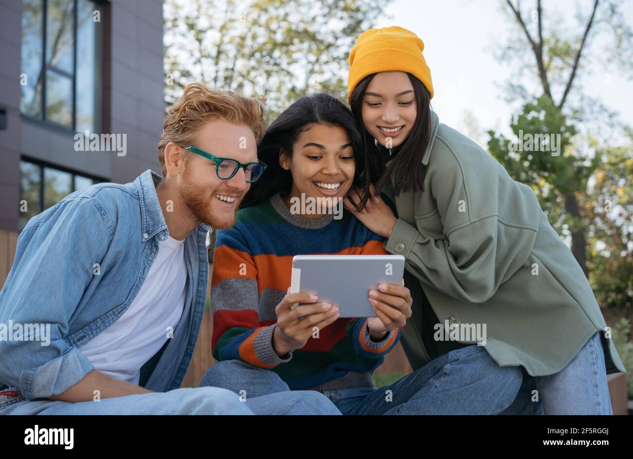 Gruppo di amici multietnici sorridenti che utilizzano tablet digitale, guardano video, fare acquisti online, stare seduti all'aperto. Studenti universitari che studiano online Foto Stock