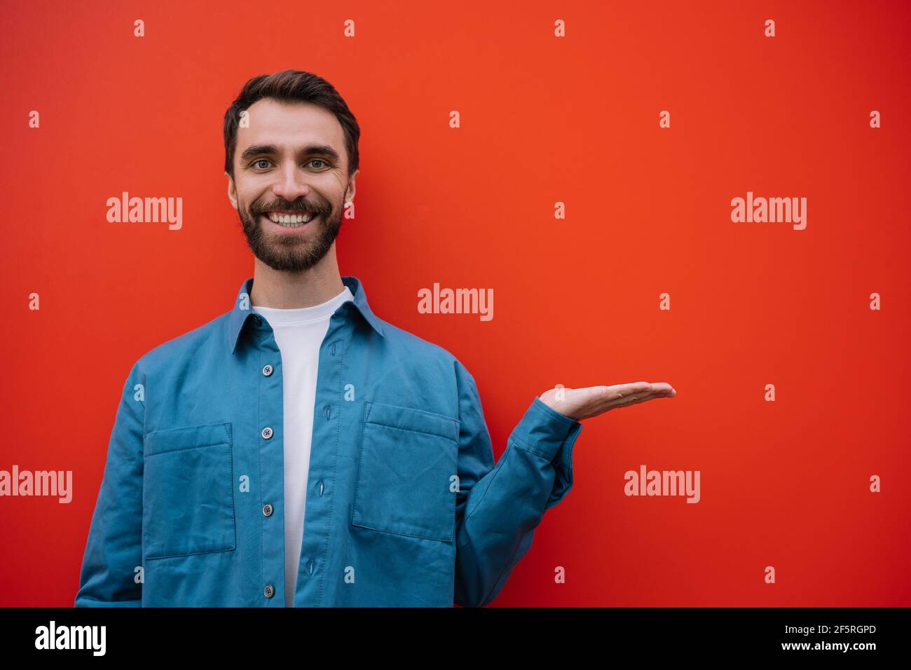 Ritratto di un giovane uomo sorridente che indossa una camicia casual che punta con la mano, isolato su sfondo rosso. Spazio di copia, concetto di pubblicità Foto Stock