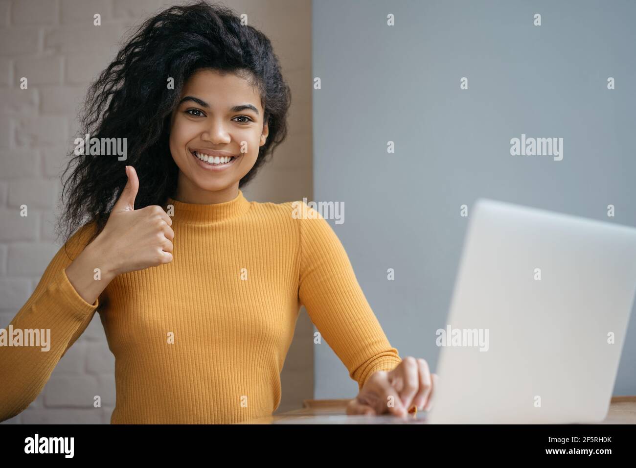 Donna afroamericana che usa un computer portatile, mostrando il pollice, guardando la fotocamera e sorridendo, lavorando da casa. Felice studente che studia online Foto Stock
