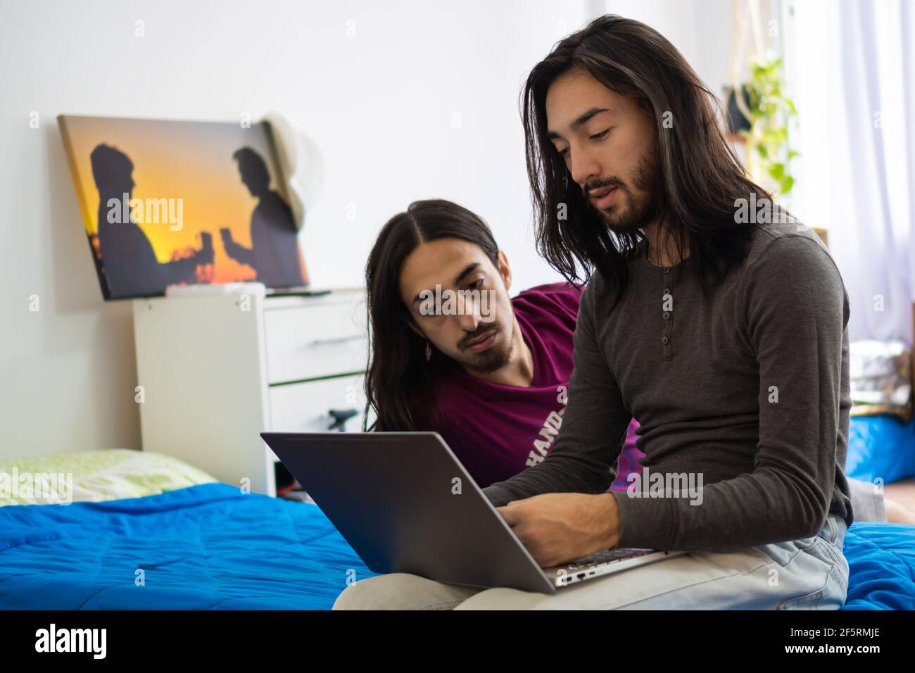 fratelli gemelli sdraiati a letto guardando il computer, 2 uomini dai capelli lunghi guardando il computer, sdraiati sul letto Foto Stock