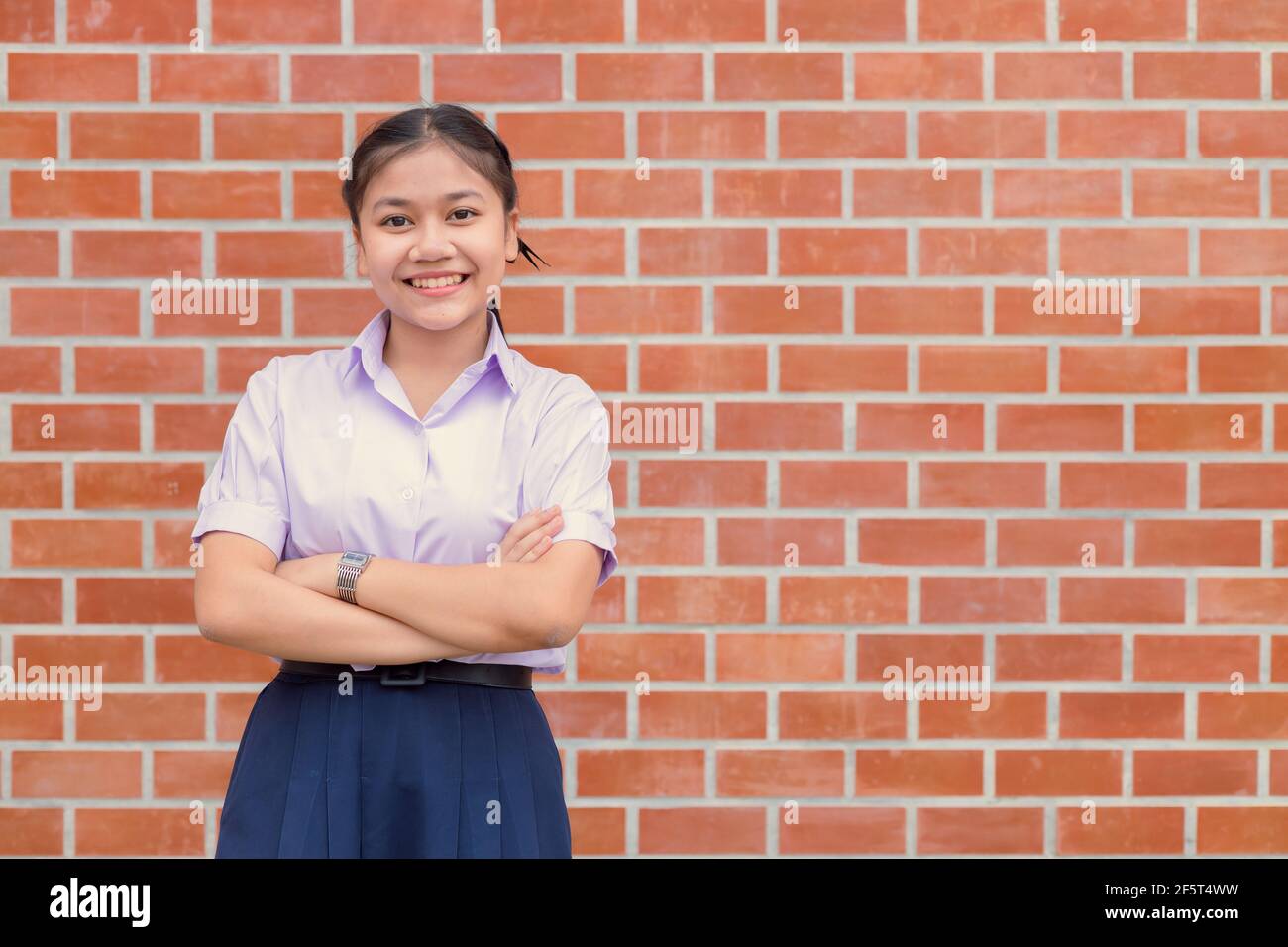 Confidente Asian Woman Student Arm incrociato uniforme sorriso felice con muro di mattoni spazio copia. Foto Stock
