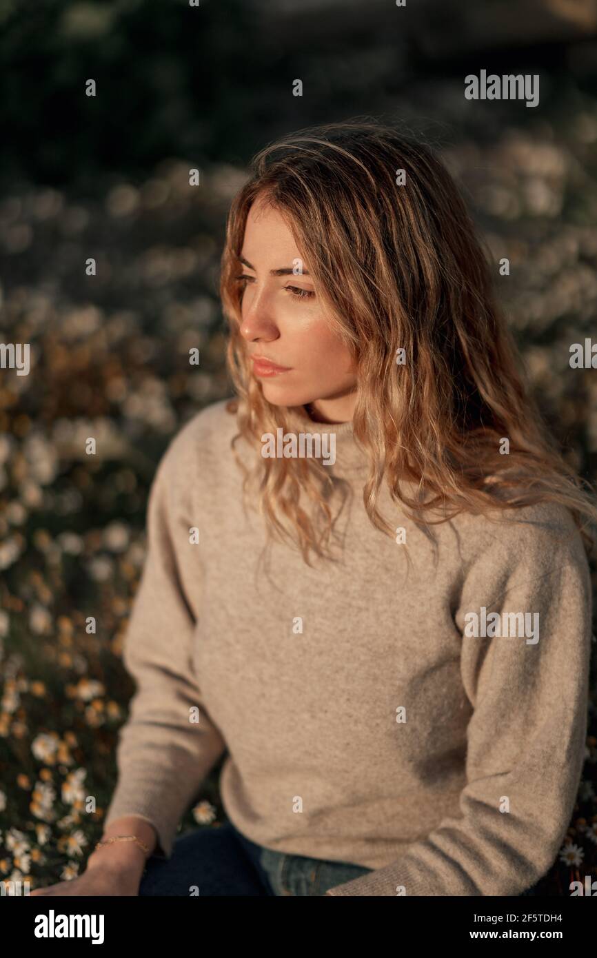 Calma contemplativa giovane viaggiatore femminile in abbigliamento casual seduto da solo su prato fiorito contro campo arato offuscato mentre ci si rilassa campagna i Foto Stock
