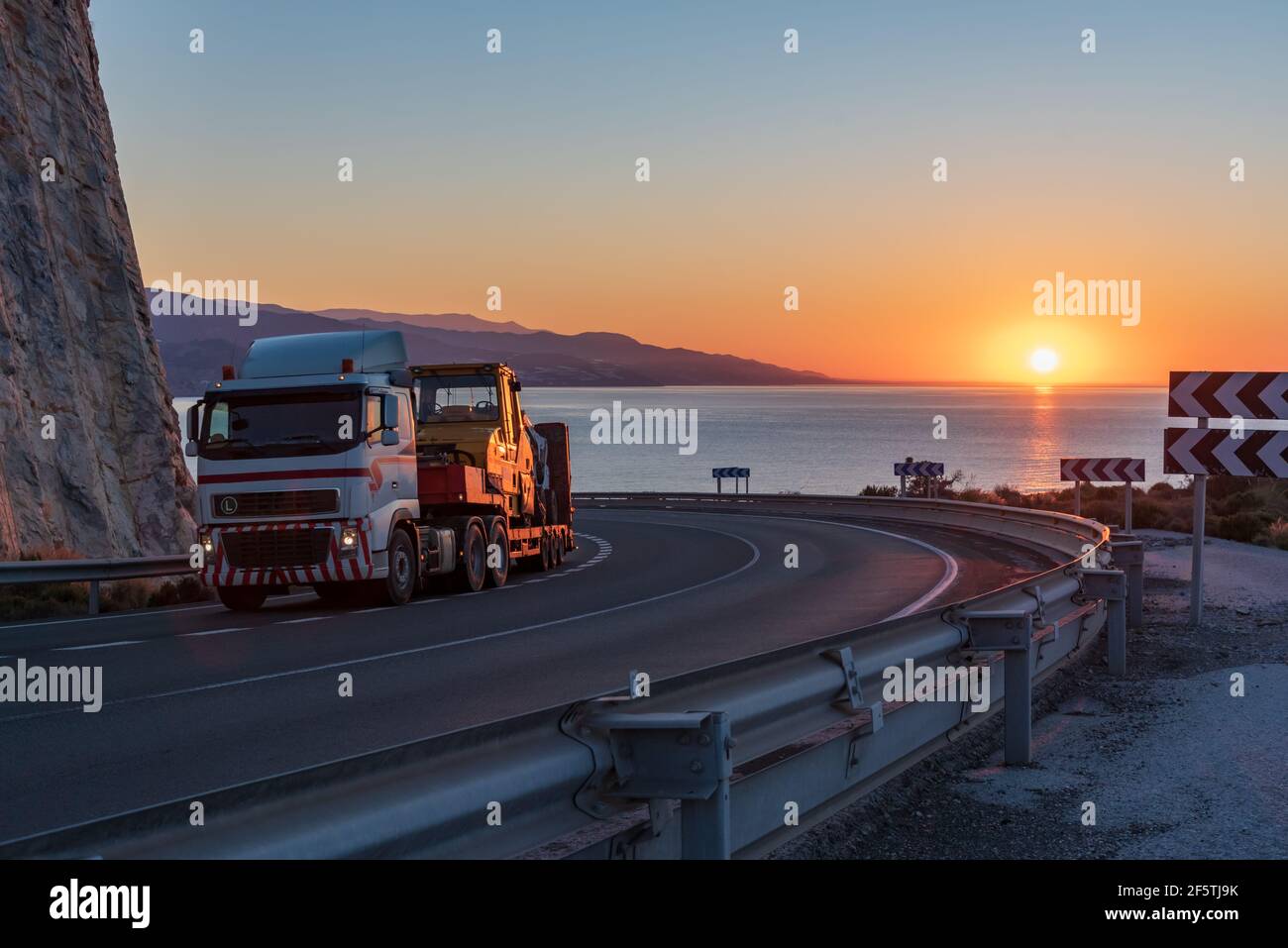 Autocarro con semirimorchio per il trasporto di macchine pesanti da costruzione su una strada di montagna con il sole all'alba sull'orizzonte sul mare. Foto Stock