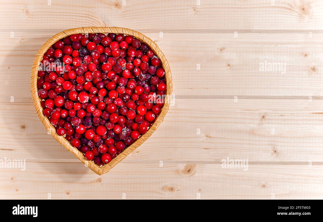 Una tavola di vimini a forma di cuore, ripiena di mirtilli rossi surgelati. Sul piano di legno chiaro a sinistra. Foto Stock