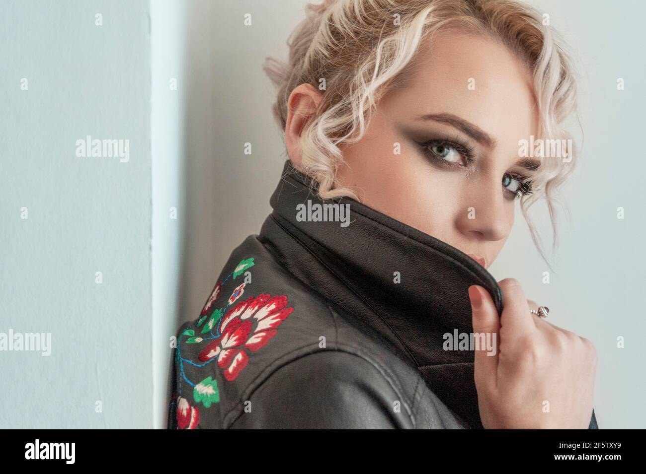 Scatto stretto di giovane donna con ombra scura dell'occhio che guarda sopra il collare della sua giacca di cuoio, appoggiandosi contro una parete interna. Foto Stock