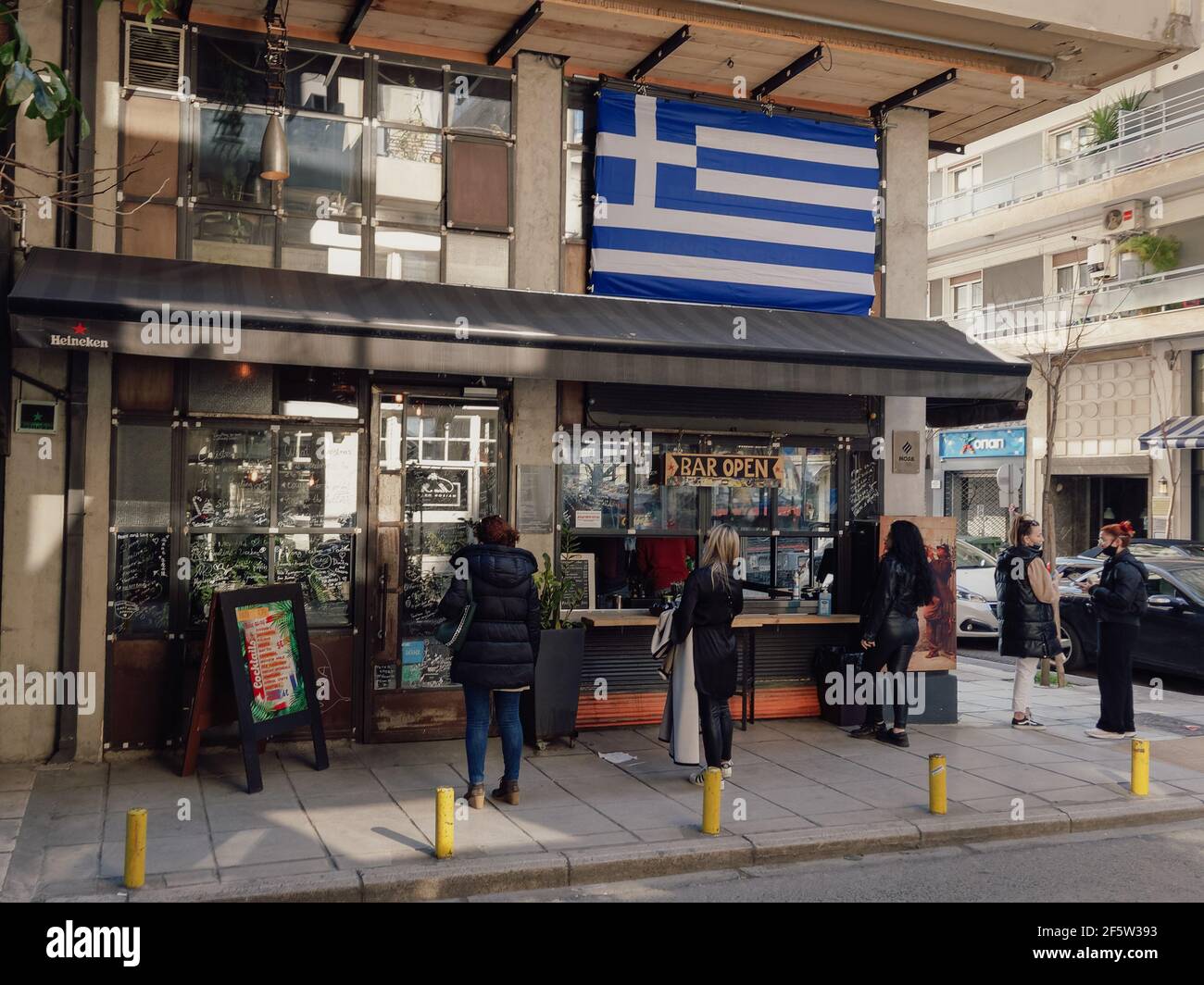 Salonicco, Grecia - Marzo 27 2021: Clienti al di fuori del banco di lavoro da take-away a causa delle restrizioni del covid-19. La folla ellenica sul marciapiede compra la propria bevanda da un negozio con una grande bandiera greca sopra. Foto Stock