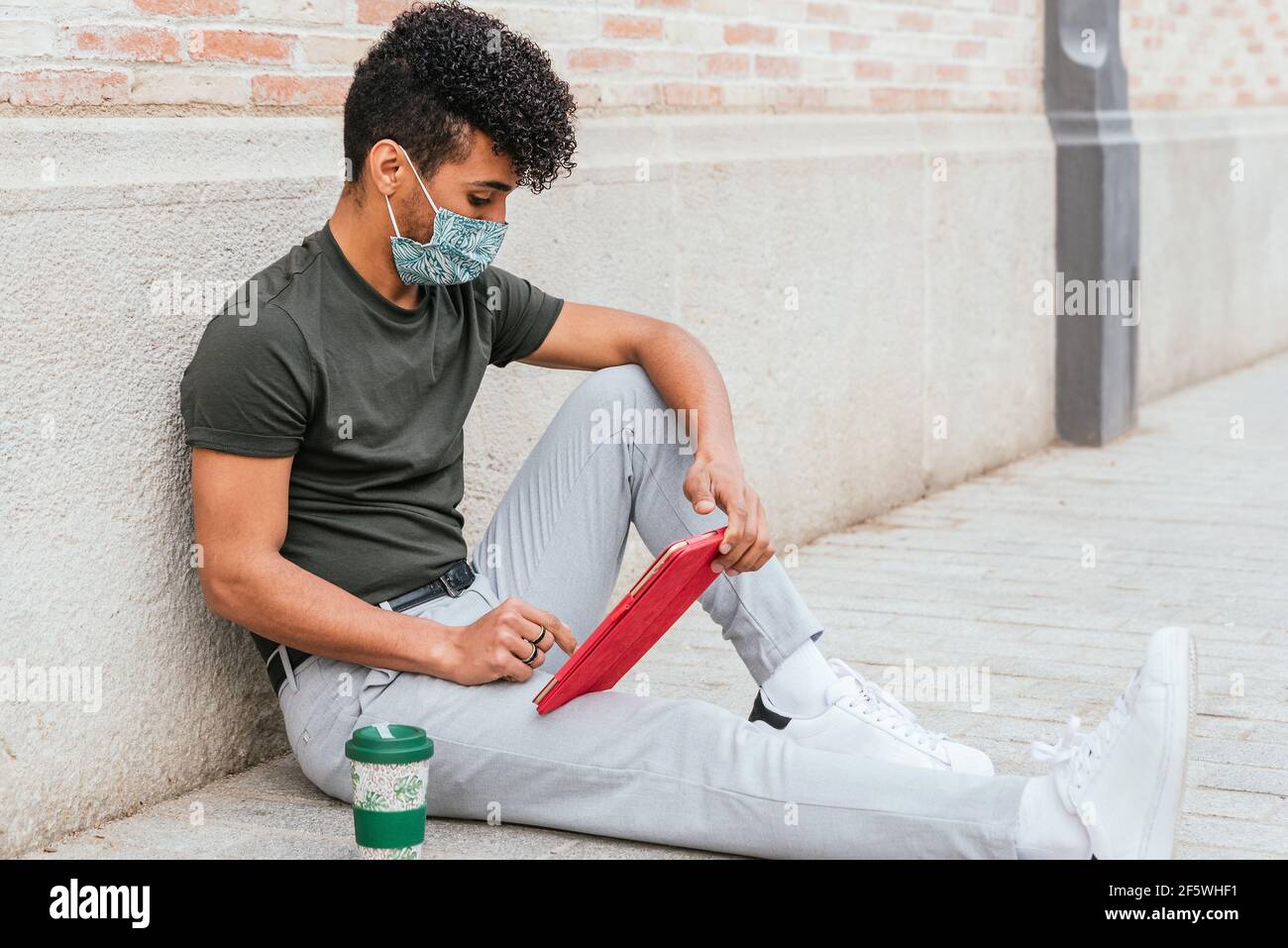 ritratto di un attraente uomo latino-americano seduto sul pavimento della strada con un tablet rosso. Si sta vestendo casual e indossa una maschera protettiva per il viso. Ha una tazza di caffè riutilizzabile vicino a lui Foto Stock