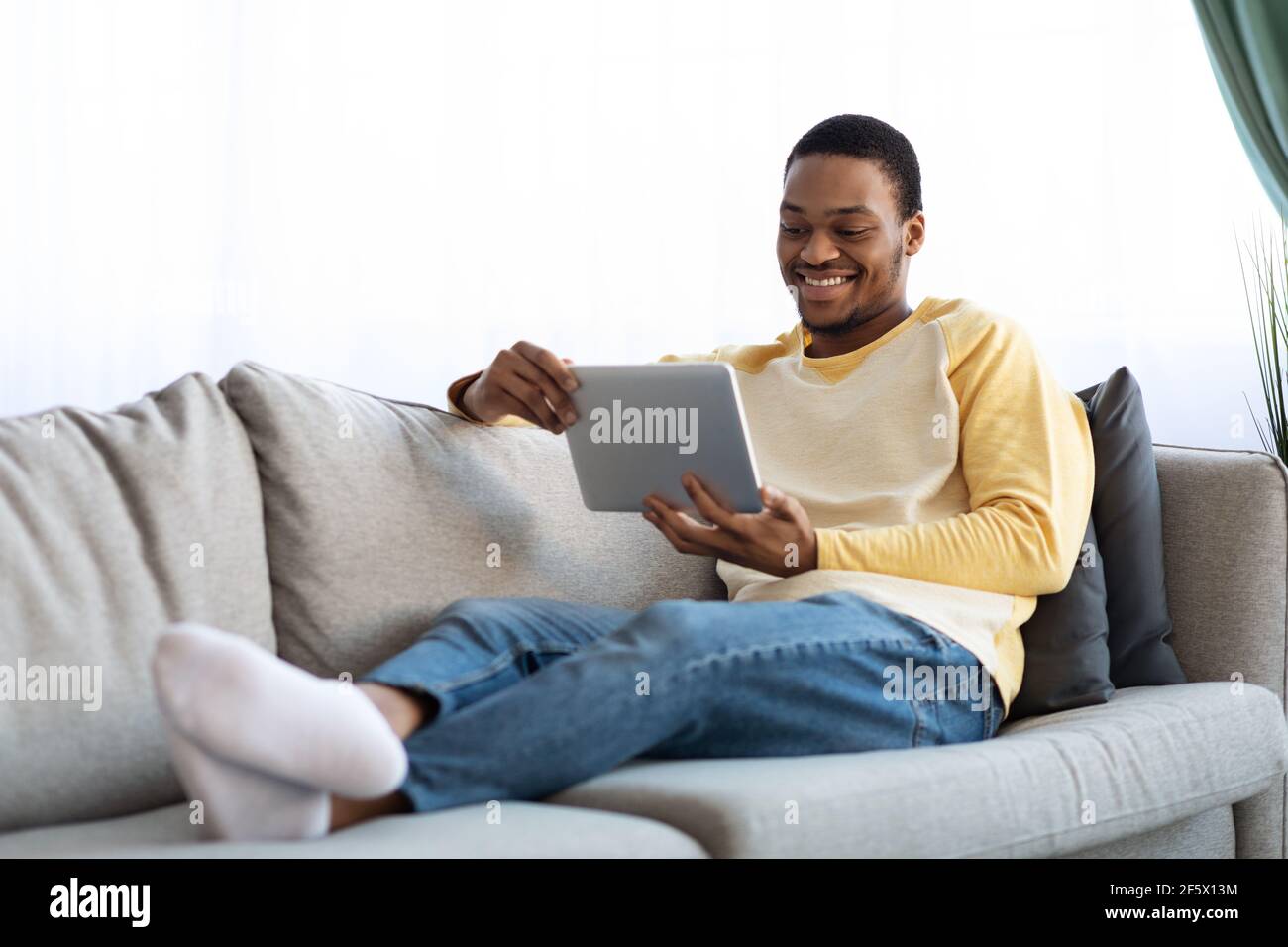 Uomo nero positivo reclinato sul divano, utilizzando un tablet digitale Foto Stock