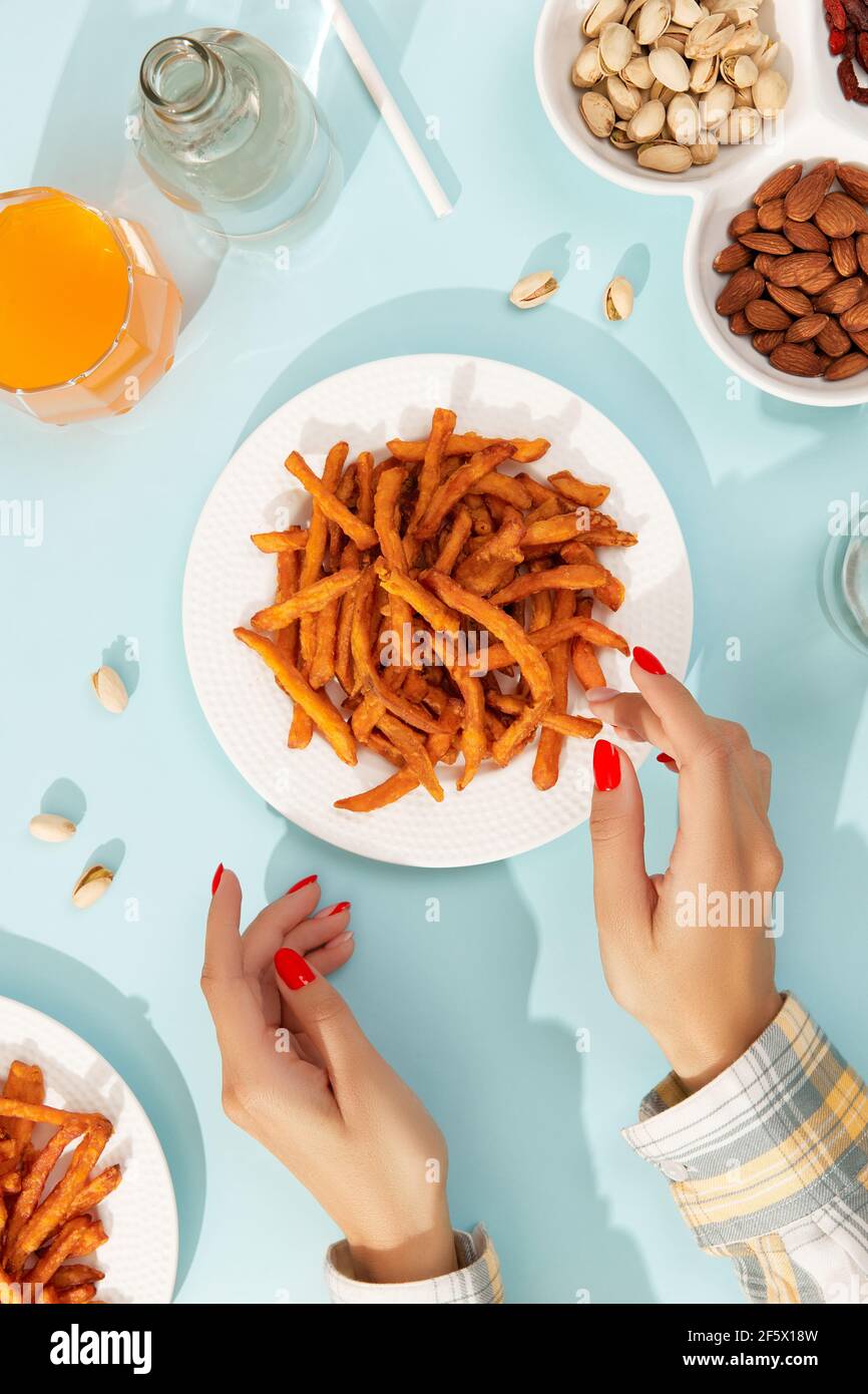 Le mani delle donne prendono le patatine dolci, le noci e le bevande su sfondo blu con le ombre lunghe in stile minimal Foto Stock
