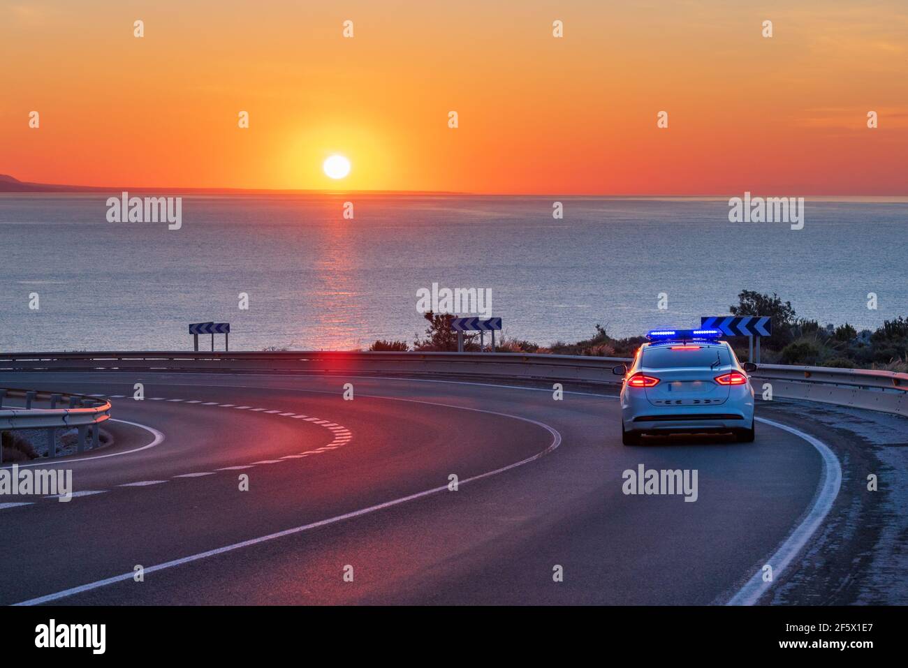 La polizia pattuglia l'auto incaricata di monitorare il traffico sulle strade, guidando su una strada di montagna con il mare e il sole all'orizzonte. Foto Stock