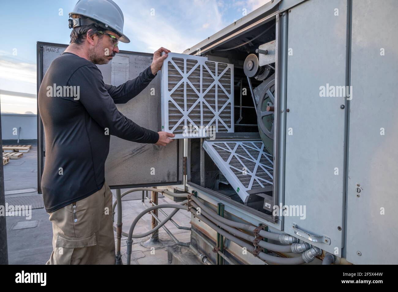 Meccanico HVAC che effettua una sostituzione del filtro dell'aria su un'unità di aria di costituzione commerciale sul tetto di un edificio. Foto Stock
