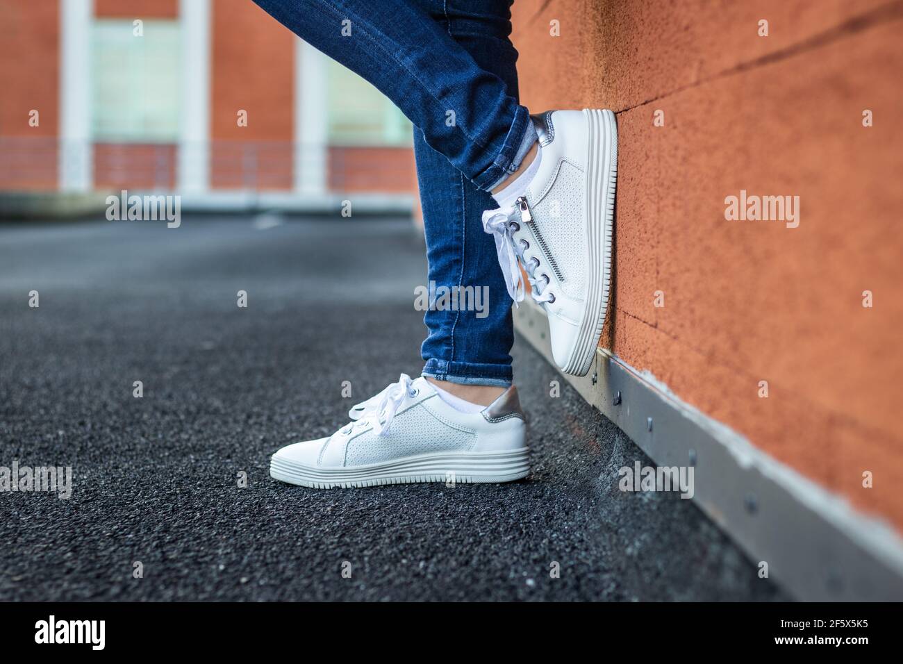 Donna con sneaker bianche in pelle a parete pendente in città, vista ravvicinata sulle gambe femminili che indossano jeans e scarpe sportive. Concetto di moda Foto Stock
