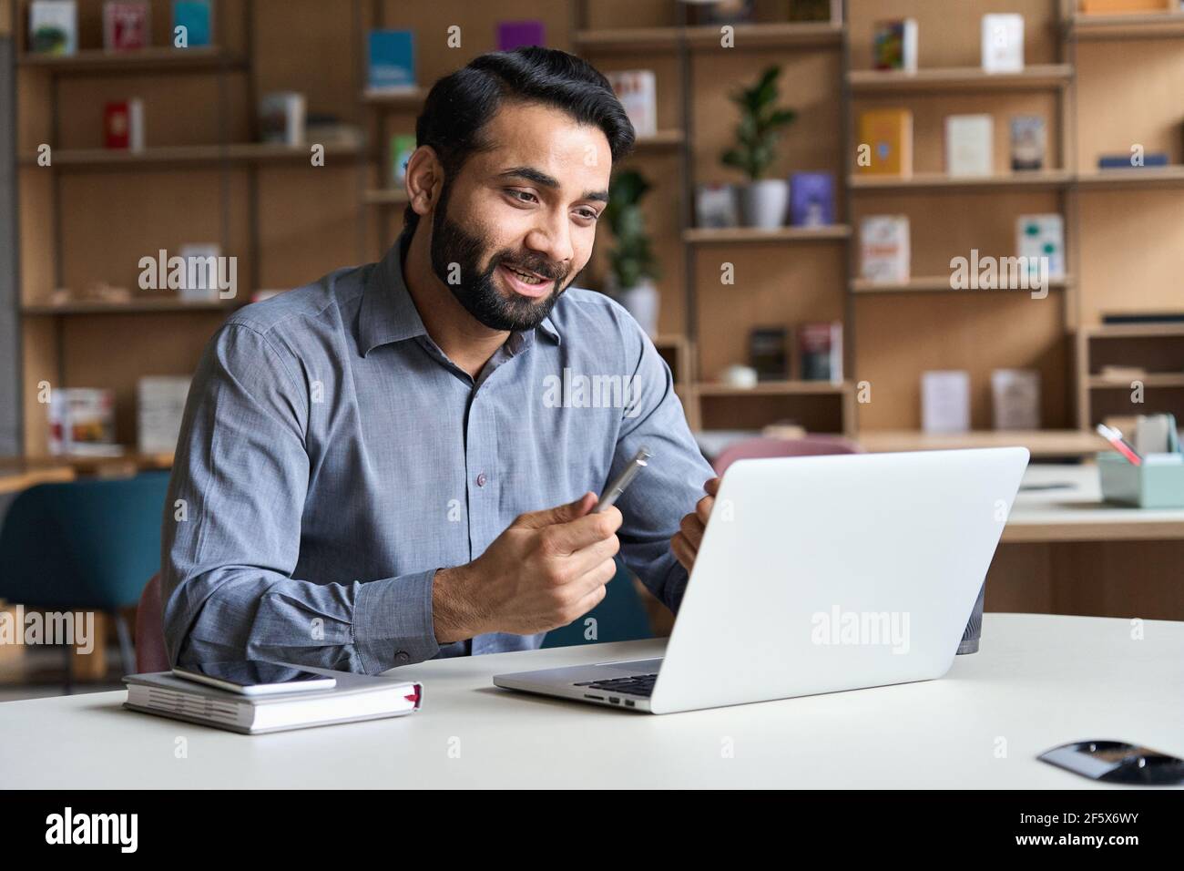 Giovane uomo d'affari indiano che parla, avendo riunione virtuale che lavora sul laptop. Foto Stock