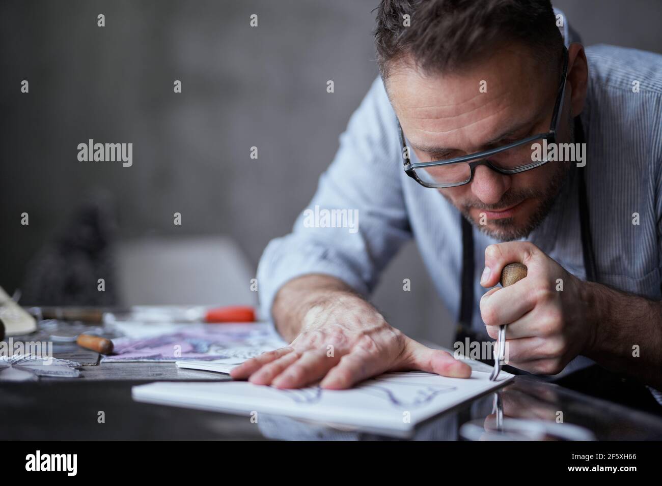 Litografia artista professionista lavoratore fatto a mano. Uomo di mezza età in grembiule e bicchieri facendo linocut utilizzando strumento di taglio. Foto di alta qualità Foto Stock