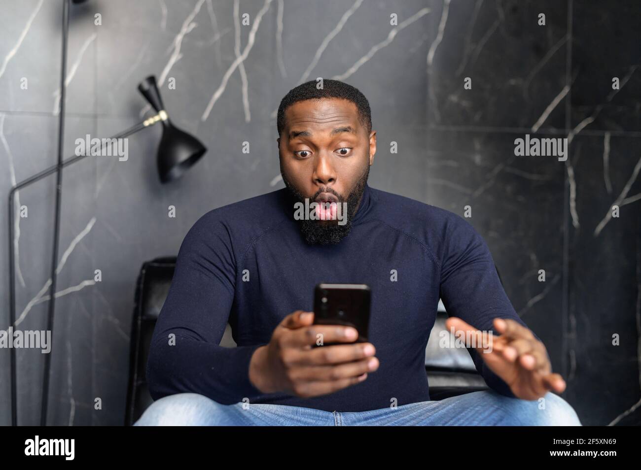 Stupito felice biracial uomo che tiene il telefono, guardando il cellulare, leggere buone notizie in sms, sedersi sulla poltrona, stupito nero ragazzo vincitore eccitato da app mobile, vincere grido con gioia, celebrare la sua vittoria Foto Stock