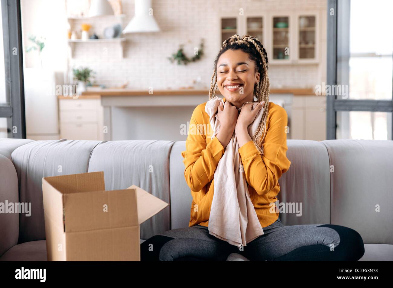 Felice soddisfatta ragazza afro-americana di stile con Dreadlock, blogger, si siede a casa, in abbigliamento casual alla moda, scompatta il pacco, abbracciando un nuovo vestito, chiude gli occhi, sorridendo, felice con un nuovo acquisto Foto Stock