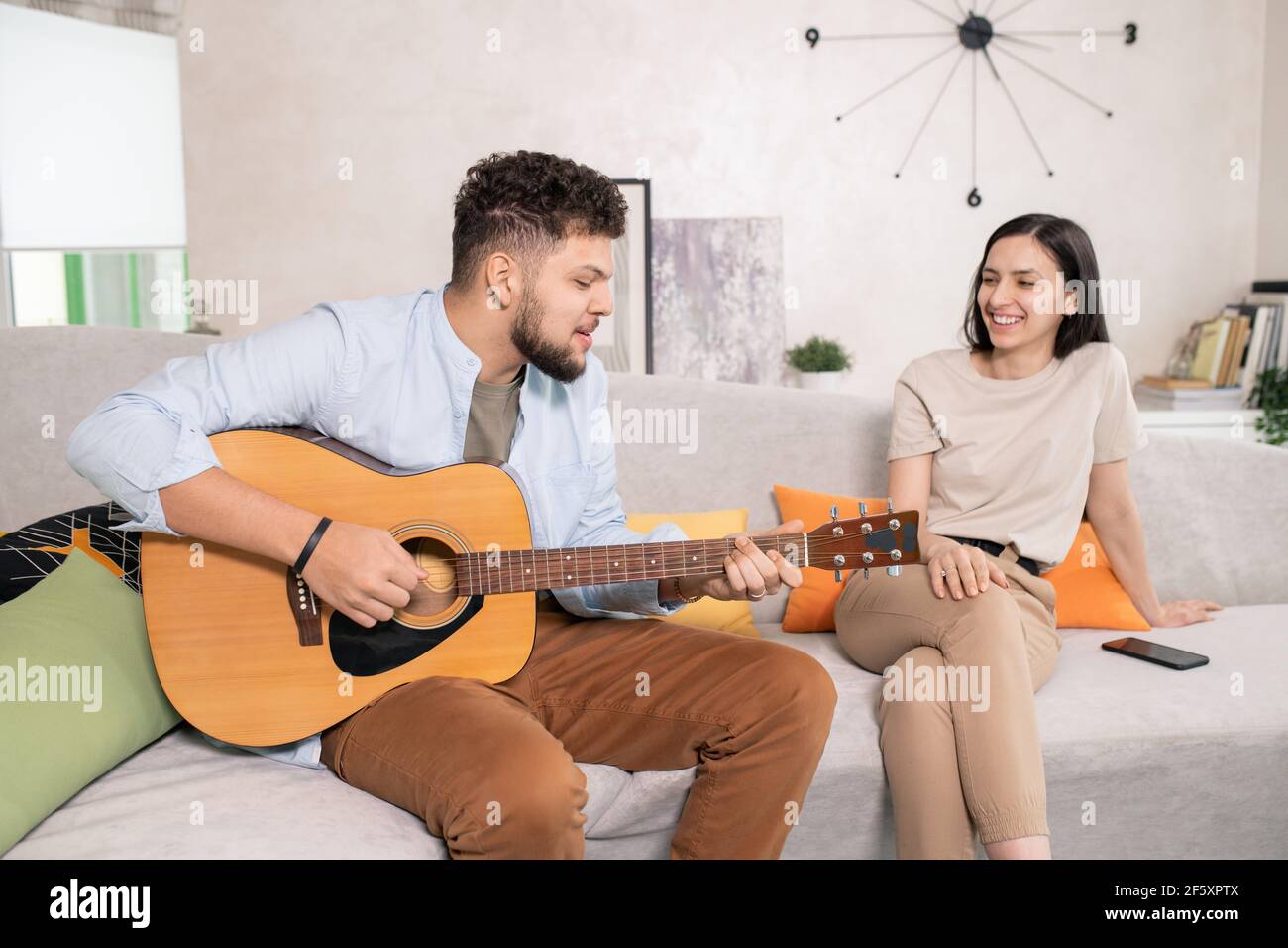 Felice giovane donna con un sorriso togoteo seduto sul divano dentro salotto e guardando suo marito suonare la chitarra e. cantare per lei Foto Stock