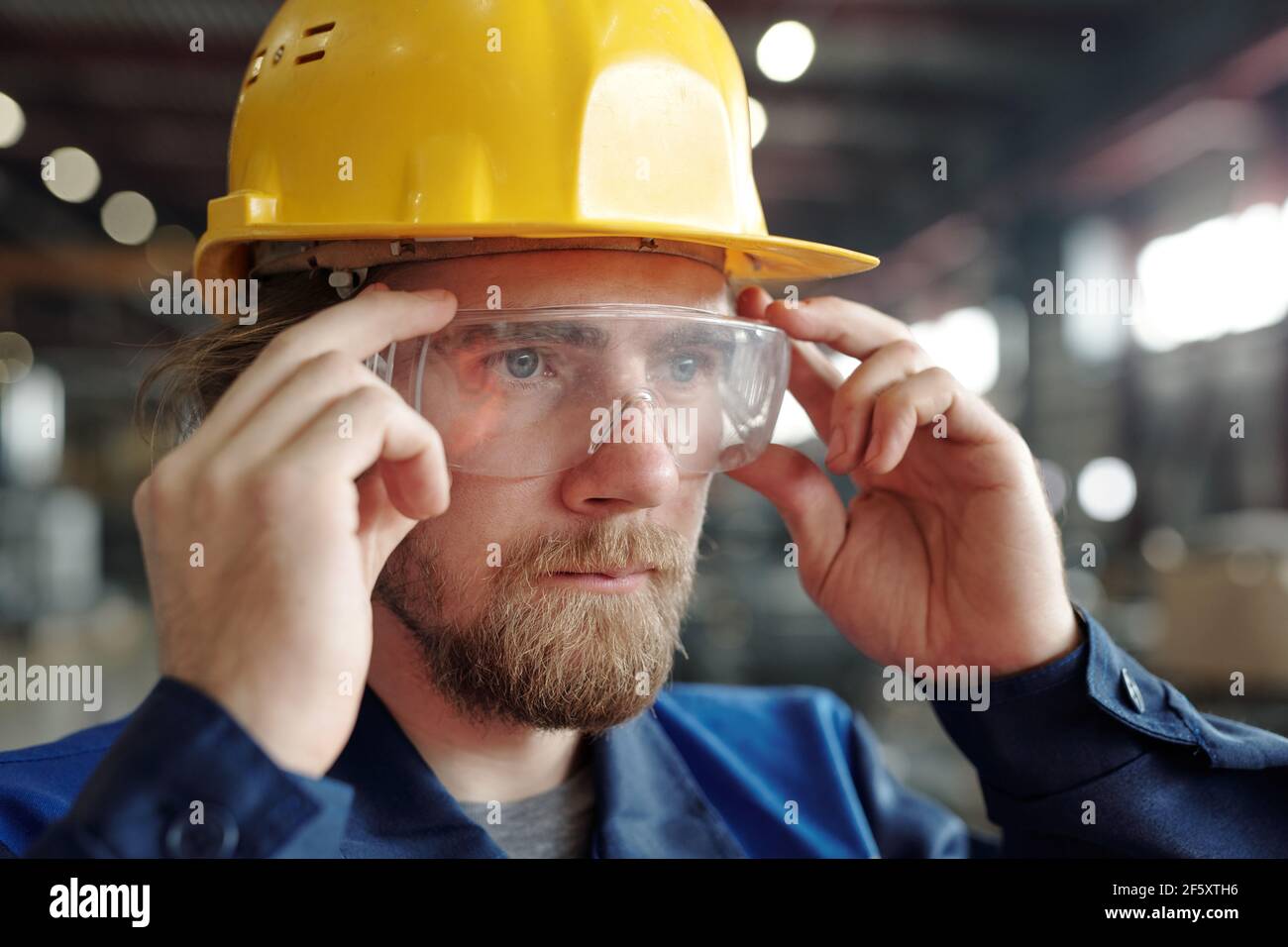 Giovane ingegnere maschile serio o lavoratore in fabbrica in casco protettivo e gli occhiali che si trovano davanti alla telecamera in un ambiente di officina Foto Stock