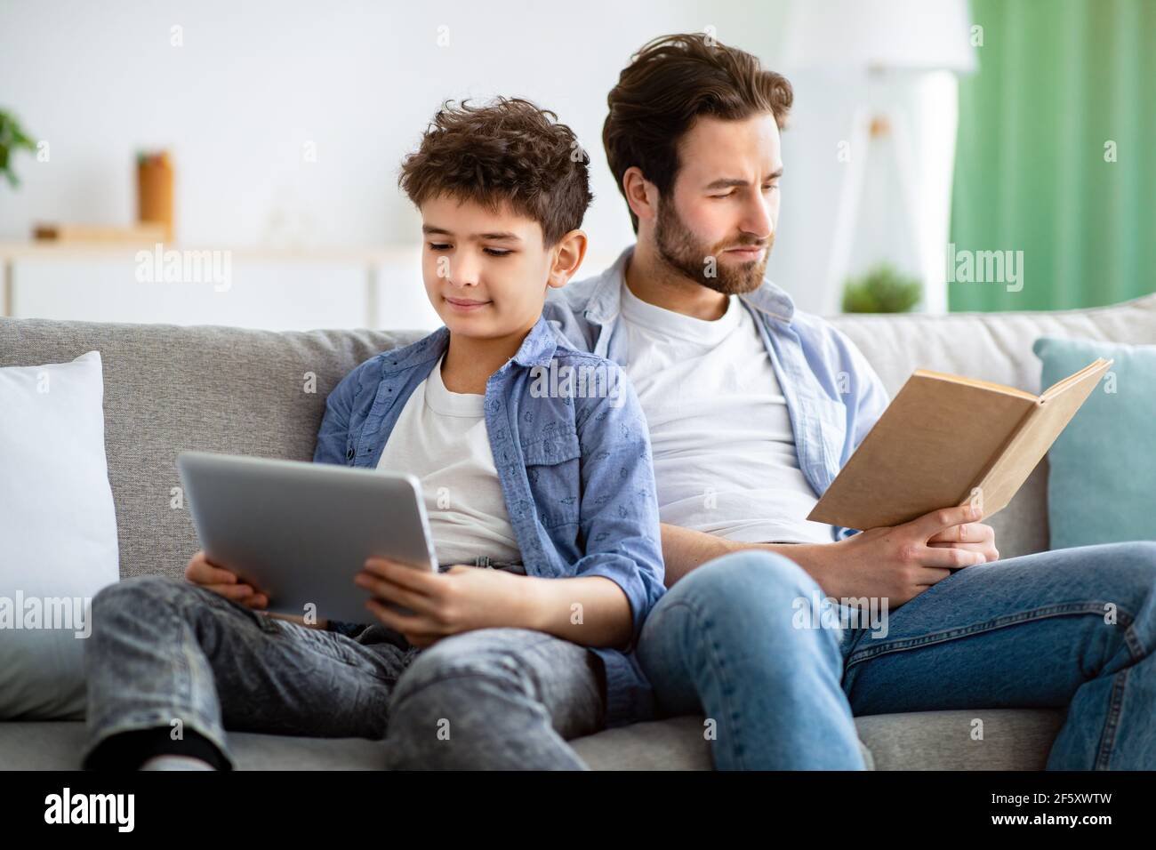 Nessuna comunicazione in famiglia. Padre leggere libro mentre suo figlio usa un tablet digitale, seduto insieme sul divano Foto Stock