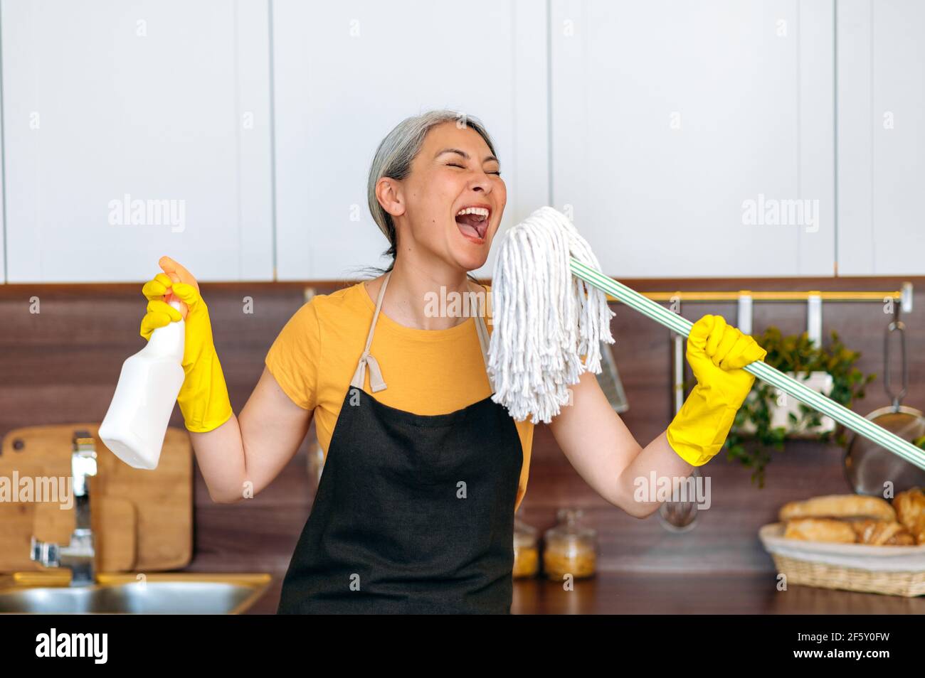Felice divertente lavorante duro maturo grigio-capelli asiatico operaio di pulizia femminile o casalinga in guanti e grembiule, prendendo una pausa, ballando in cucina con detergente e mop, cantando la sua canzone preferita Foto Stock
