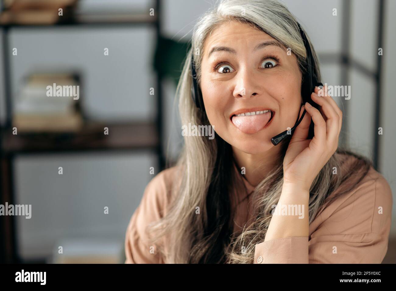 Ritratto di una donna asiatica felice e sciocco con capelli grigi maturi in cuffia, dipendente del call center, manager, freelance, guardando la fotocamera, imbrogliare, mostrare la lingua, divertirsi, faccia divertente Foto Stock