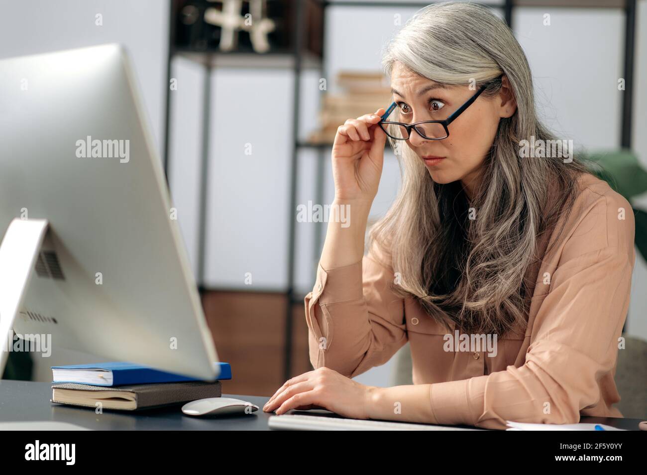 Scioccato entusiasta influente maturo grigio-capelli asiatici, donna d'affari, manager, è seduto al computer, ha ricevuto un messaggio inaspettato, guarda sorpreso lo schermo del computer, toglie gli occhiali Foto Stock