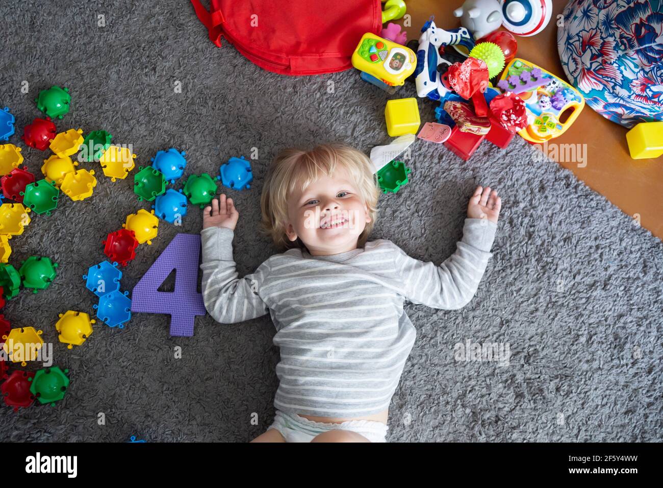 Il bambino biondo felice si trova sul pavimento con i lotti giocattoli Foto Stock