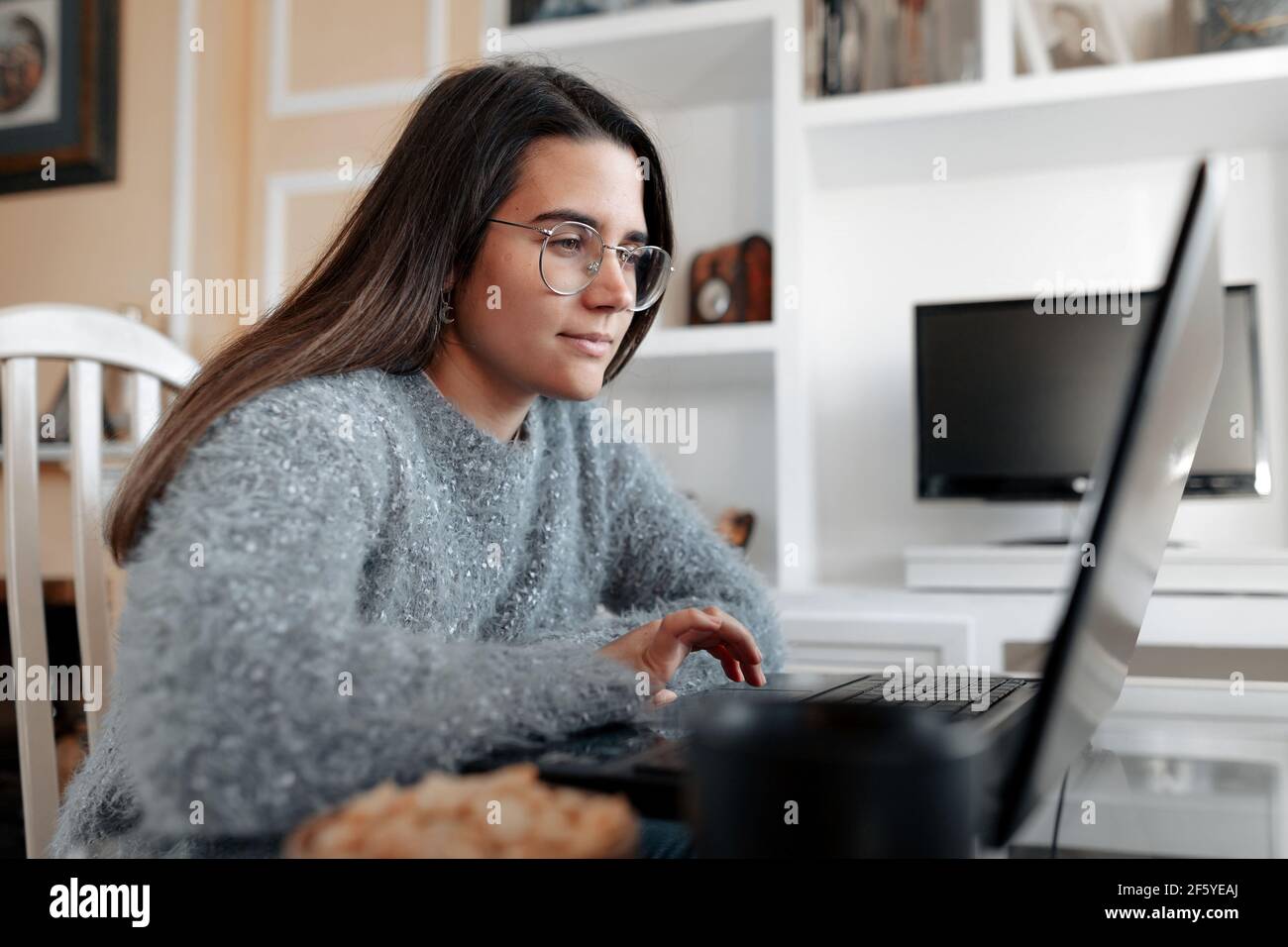 Giovane donna allegra che indossa un maglione a casa sua è seduta nel suo soggiorno mentre naviga in Internet nel computer portatile. Foto Stock