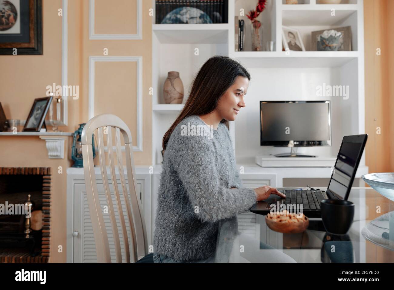 Giovane donna allegra che indossa un maglione a casa sua è seduta nel suo soggiorno mentre naviga in Internet nel computer portatile. Foto Stock