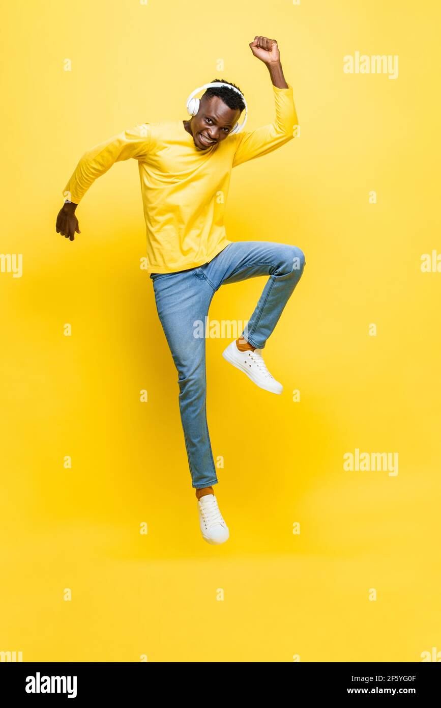 Felice giovane africano uomo che indossa le cuffie ascoltare la musica e. saltando con mano in su in giallo isolato studio sfondo Foto Stock
