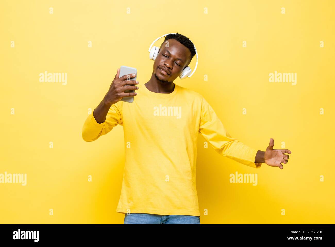 Un giovane africano che indossa le cuffie per ascoltare la musica online dal telefono cellulare in uno sfondo giallo isolato studio Foto Stock