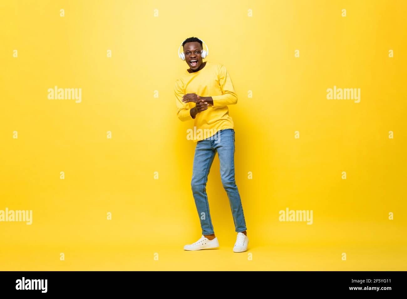 Giovane uomo africano felice che indossa le cuffie ascoltando la musica e. danza in uno sfondo giallo isolato studio Foto Stock