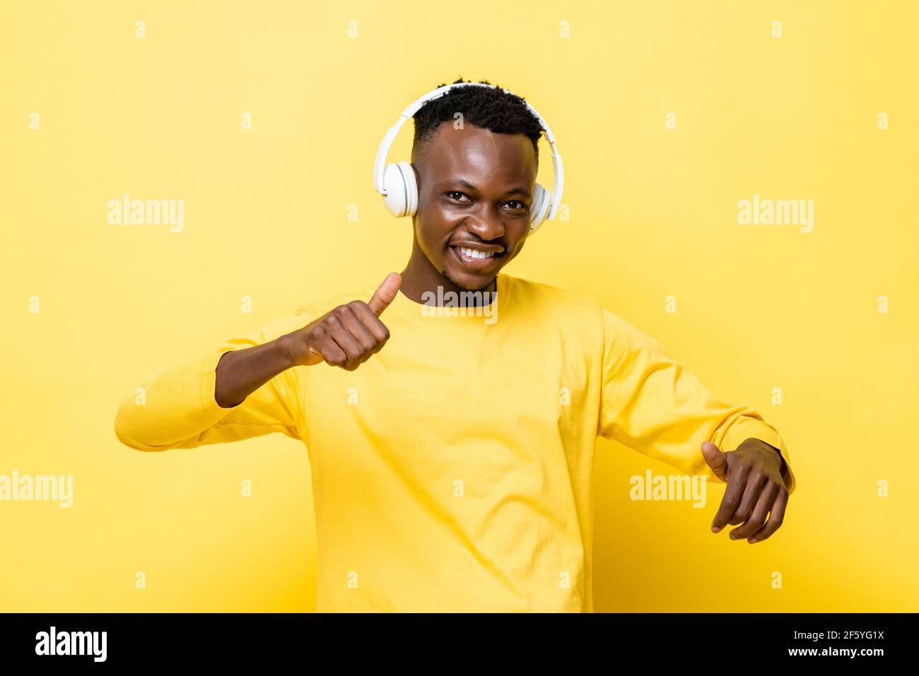 Felice sorridente giovane africano in abbigliamento casual godendo l'ascolto alla musica con le cuffie e balli sopra la parete gialla dello studio sfondo Foto Stock