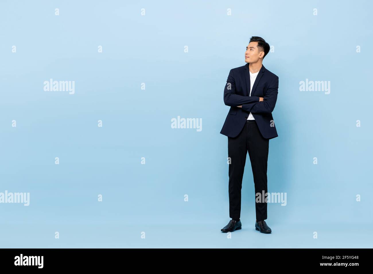 Ritratto a lunghezza intera di giovane bell'uomo d'affari del sud-est asiatico con braccia incrociate guardando verso l'alto lateralmente per copiare spazio sulla luce sfondo blu studio Foto Stock