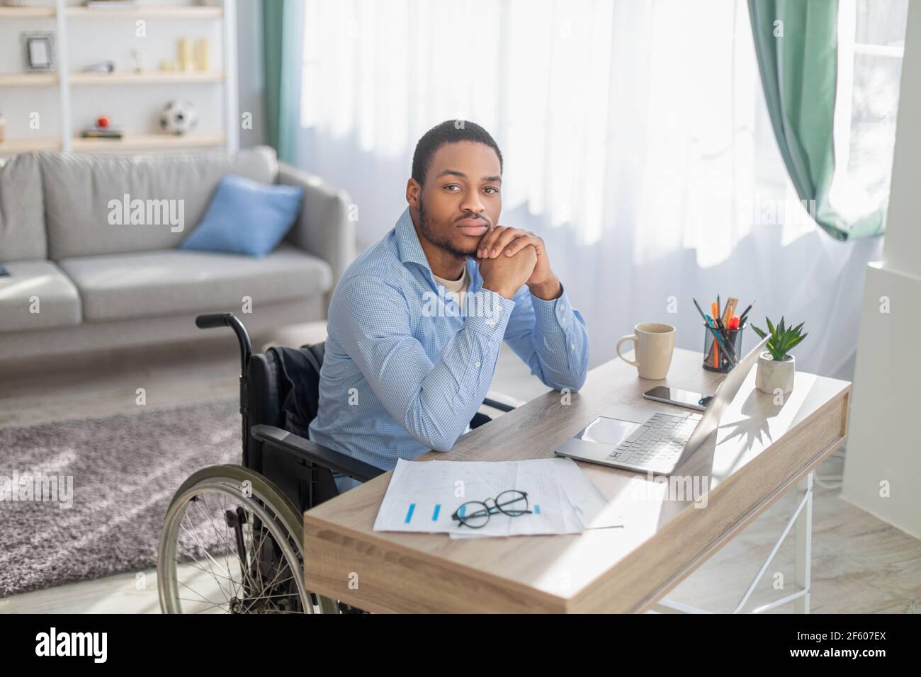 Lavoro online per disabili. Serio ragazzo nero in sedia a rotelle seduto alla scrivania con il computer portatile, guardando la fotocamera, in interni Foto Stock