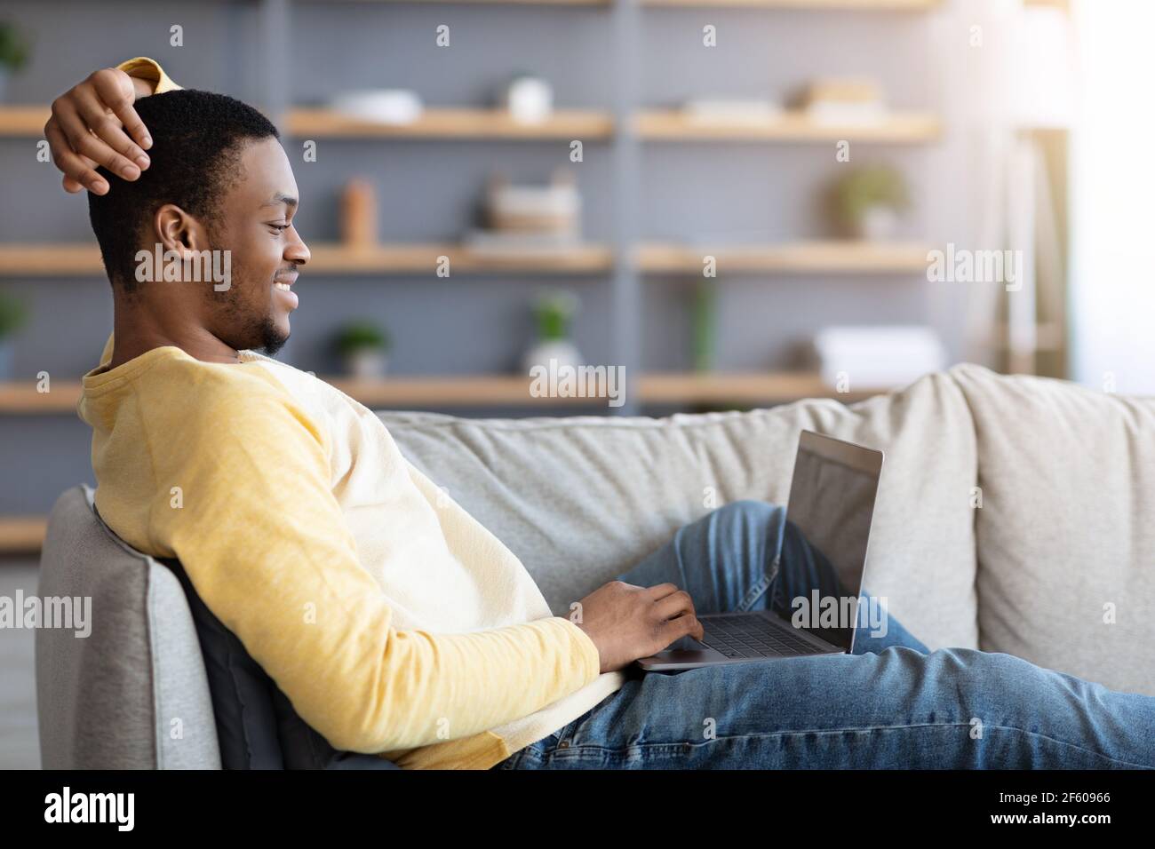Felice americano aficano ragazzo che riposa sul divano, usando il laptop Foto Stock